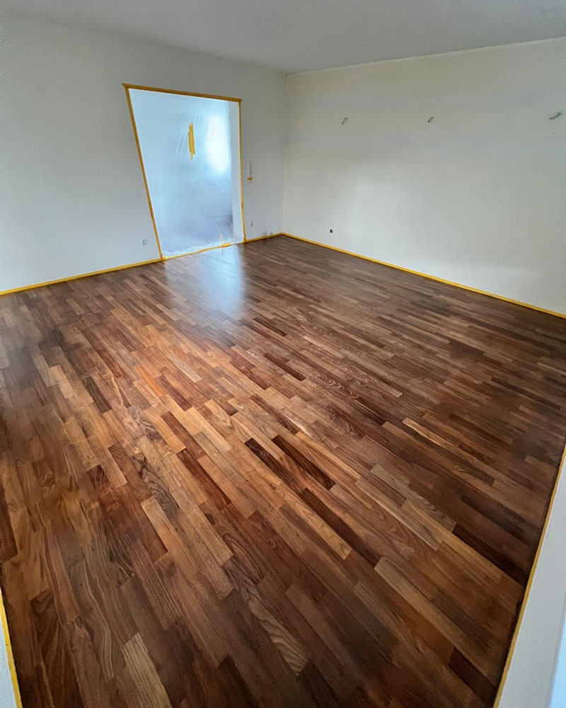 Ein frisch verlegter Holzfußboden mit warmen, natürlichen Farbtönen erstreckt sich über einen leeren Raum. Die Wände sind vorbereitet und mit Abdeckfolie sowie gelbem Klebeband geschützt, was auf laufende Renovierungsarbeiten hinweist. Die Szene zeigt die hochwertige Verlegung eines neuen Bodenbelags sowie die sorgfältige Vorbereitung des Raums für weitere Arbeitsschritte.