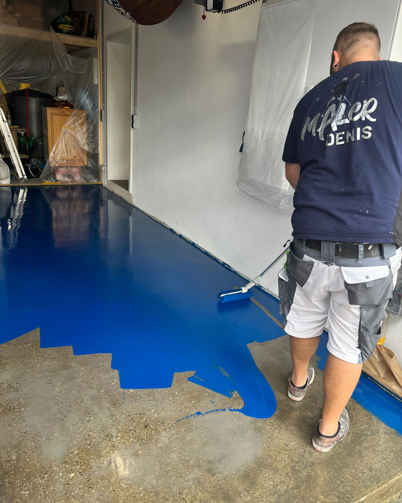 Ein Maler trägt mit einer Farbrolle eine kräftig blaue Bodenbeschichtung auf den Garagenboden auf. Der bereits beschichtete Bereich glänzt, während der rohe Betonboden im Vordergrund noch unbehandelt ist. Im Hintergrund sind abgedeckte Möbel und Werkzeuge zu sehen. Die Szene zeigt den professionellen Auftrag einer widerstandsfähigen Bodenbeschichtung im Innenbereich.