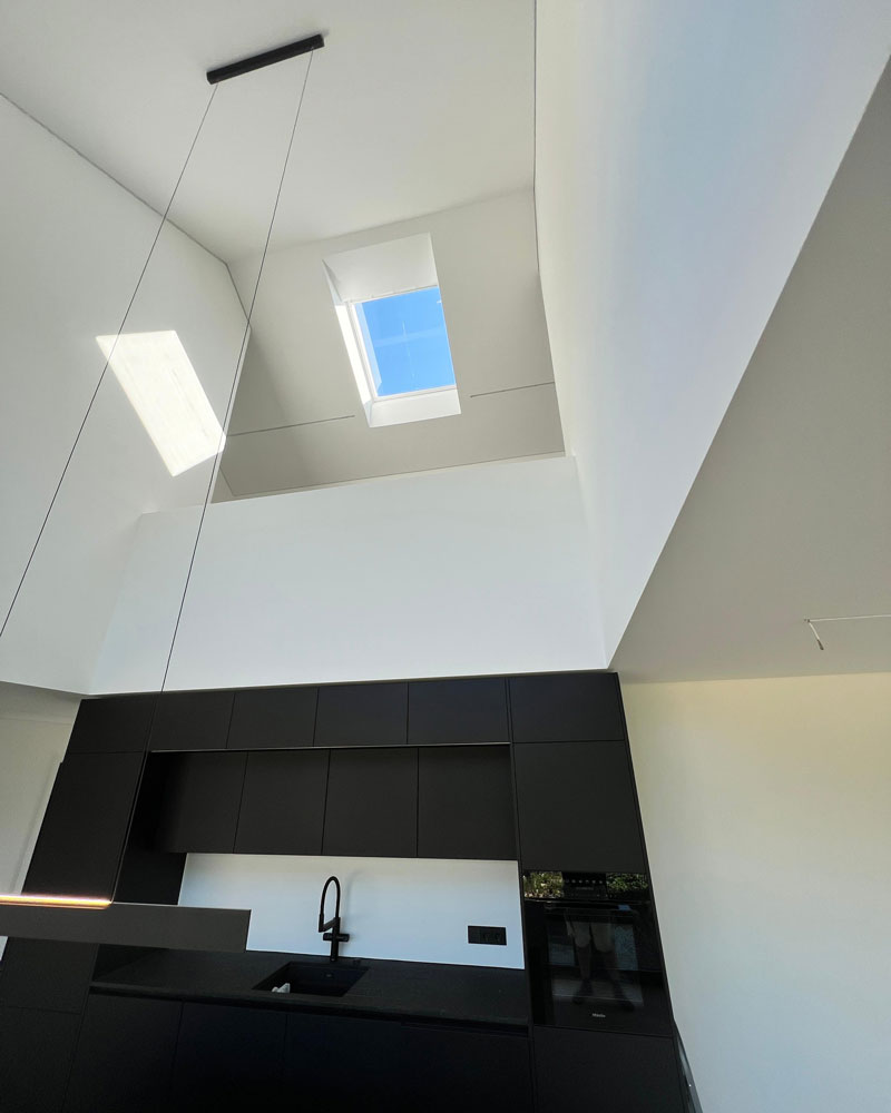 Eine moderne Küche mit schwarzen Einbaumöbeln und klaren Linien, darüber eine hohe weiße Decke mit großem Dachfenster. Durch das Fenster fällt viel Tageslicht ein, wodurch die glatten Wand- und Deckenflächen besonders sauber und präzise wirken. Die Szene zeigt hochwertige Maler- und Spachtelarbeiten in einem architektonisch anspruchsvollen Innenraum.