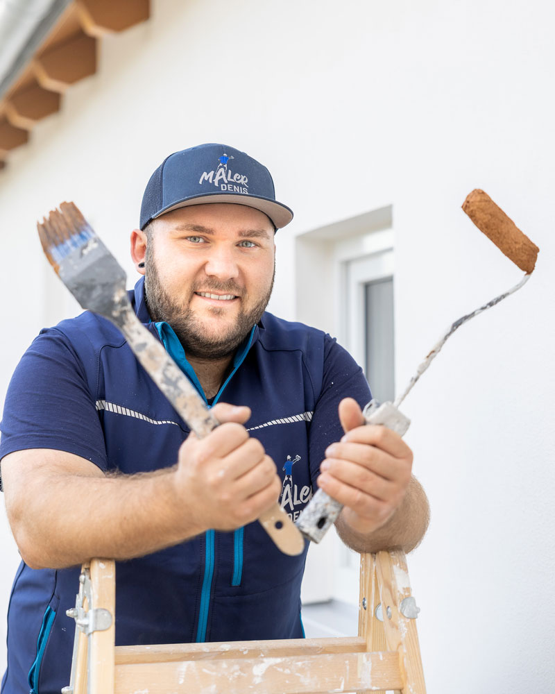 Ein Maler steht auf einer Stehleiter und hält einen Pinsel sowie eine Farbrolle in den Händen. Er trägt Arbeitskleidung und eine Kappe mit Firmenlogo. Im Hintergrund ist eine frisch gestrichene weiße Fassade zu sehen. Die Szene vermittelt professionelle Malerarbeiten und den persönlichen Kontakt zum Handwerker.