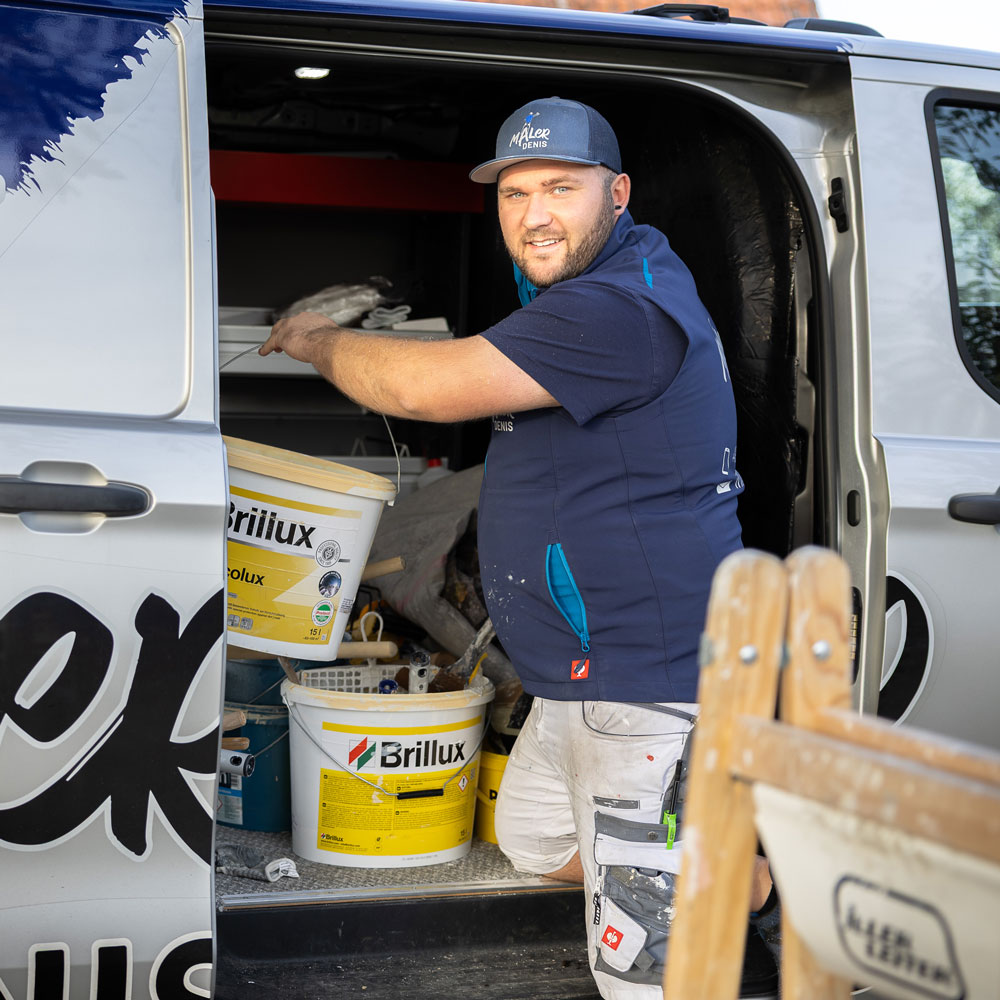 Ein Maler entnimmt Farbeimer und Werkzeug aus dem geöffneten Firmenfahrzeug. Er trägt Arbeitskleidung und eine Kappe mit Firmenlogo. Im Inneren des Transporters sind verschiedene Materialien und Utensilien für Malerarbeiten zu erkennen. Die Szene zeigt die Vorbereitung auf einen Einsatz sowie die professionelle Ausstattung eines modernen Malerbetriebs.