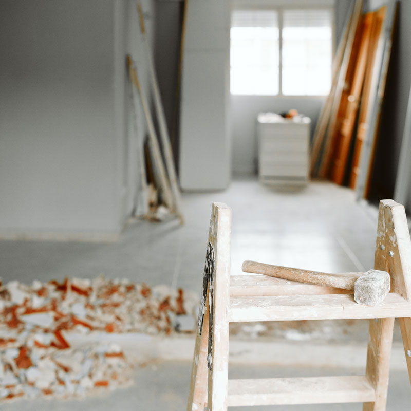 Eine Holzleiter mit einem Handhammer steht im Vordergrund eines Raums, der sich in einer umfassenden Renovierungs- oder Sanierungsphase befindet. Auf dem Boden liegen Bauschutt und herausgebrochene Wandteile, während im Hintergrund Baumaterialien und Werkzeuge bereitstehen. Die Szene zeigt typische Abläufe bei Umbauarbeiten im Innenbereich – vom Entfernen alter Strukturen bis zur Vorbereitung für neue Installationen.