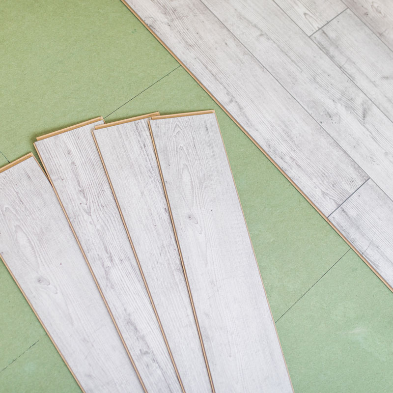 Mehrere Laminatdielen in heller Holzoptik liegen bereit zur Verlegung auf einer grünen Trittschalldämmung. Rechts im Bild ist bereits ein Teil des verlegten Bodens sichtbar. Die Szene zeigt die typischen Schritte bei der Bodenmontage – von der Vorbereitung mit Unterlage bis zum passgenauen Verbinden der einzelnen Paneele.